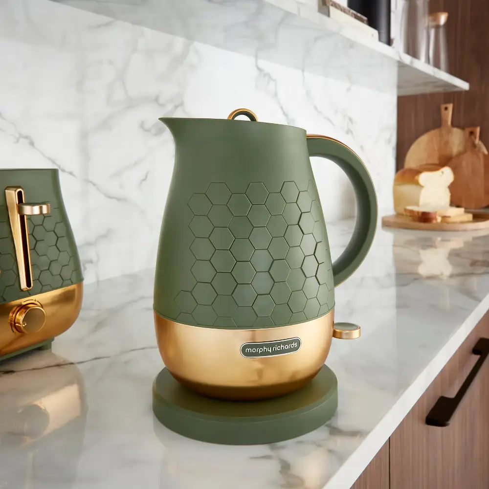 Morphy Richards Cassini 1.7 litre jug kettle in olivine green with honeycomb pattern detail and gold accents, displayed on a marble kitchen counter with matching green toaster and wooden accessories in the background