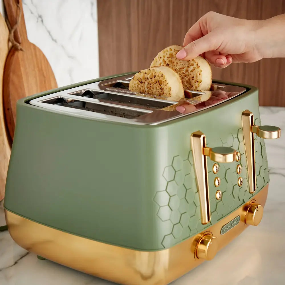Morphy Richards Cassini 4 slice toaster in sage green and gold with two toasted seeded crackers being removed from the slots, displayed on a marble countertop with wooden and brown curtain background
