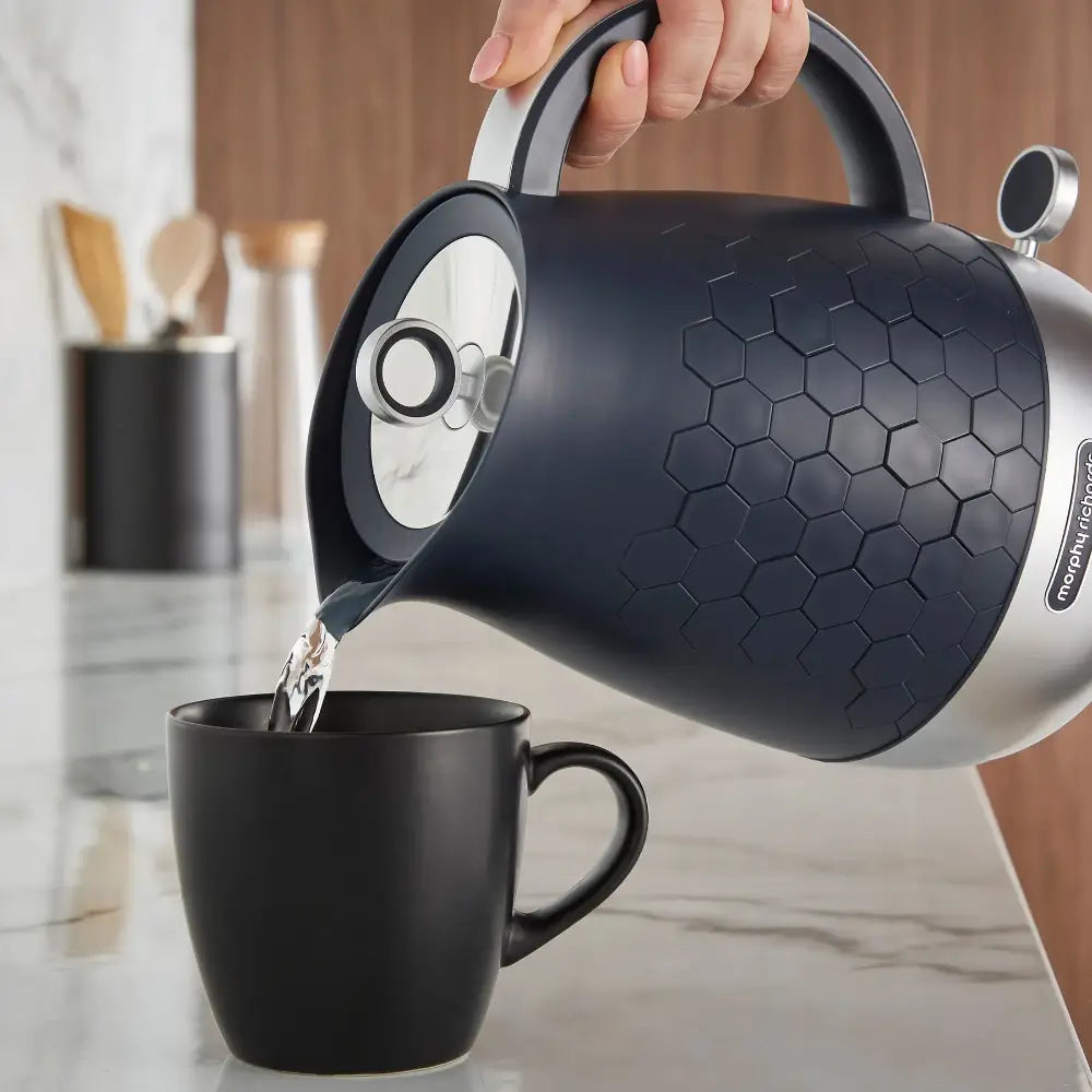 Black Morphy Richards Cassini jug kettle with hexagonal pattern design pouring hot water into a black matte mug in a modern kitchen setting