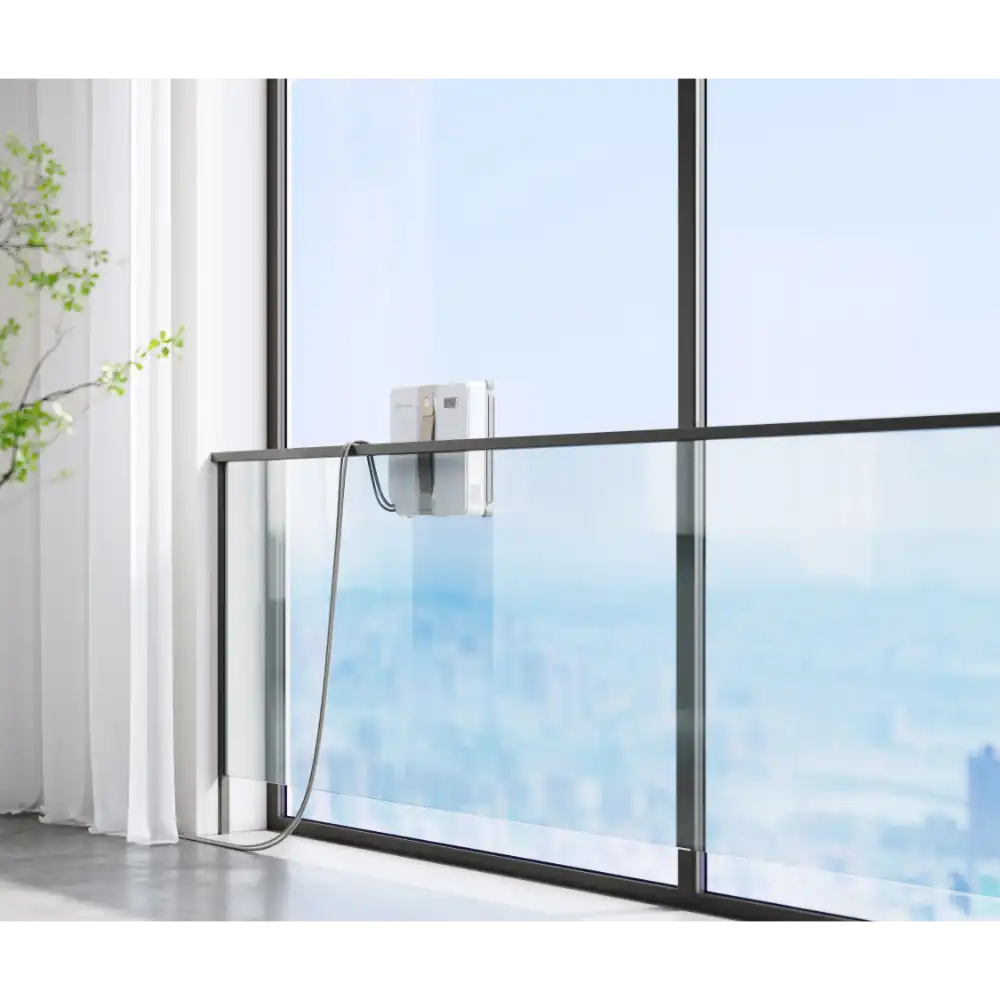 Ecovacs Winbot Mini beige robot window vacuum mounted on modern floor-to-ceiling window with black frame, cleaning glass pane with city skyline view, white curtains and green plant visible to the left