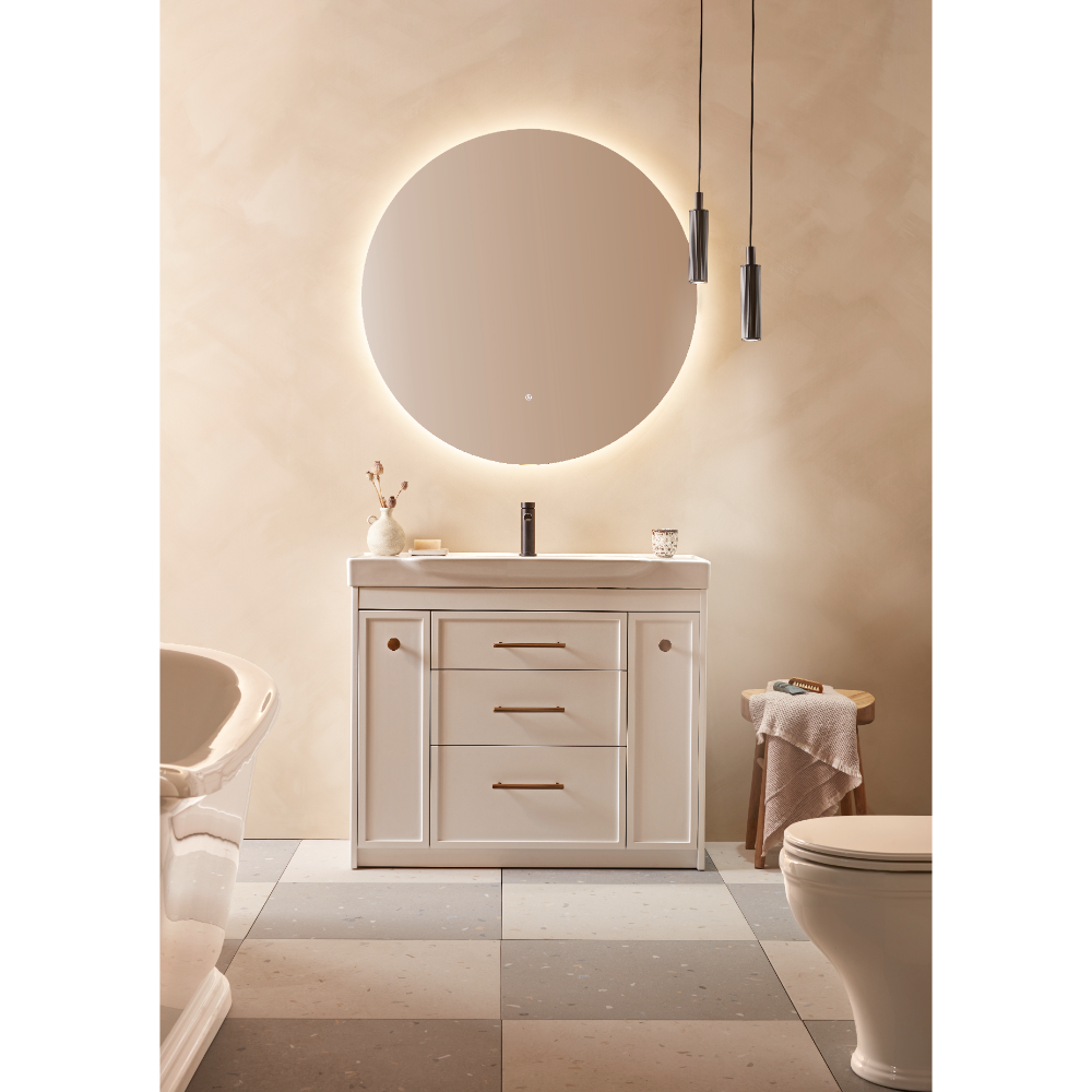 Modern bathroom vanity unit in matt white finish with integrated basin, three central drawers with bronze handles, flanked by cupboard doors, positioned beneath an illuminated circular mirror with warm LED backlighting, styled in a contemporary luxury bathroom setting with beige and cream tones