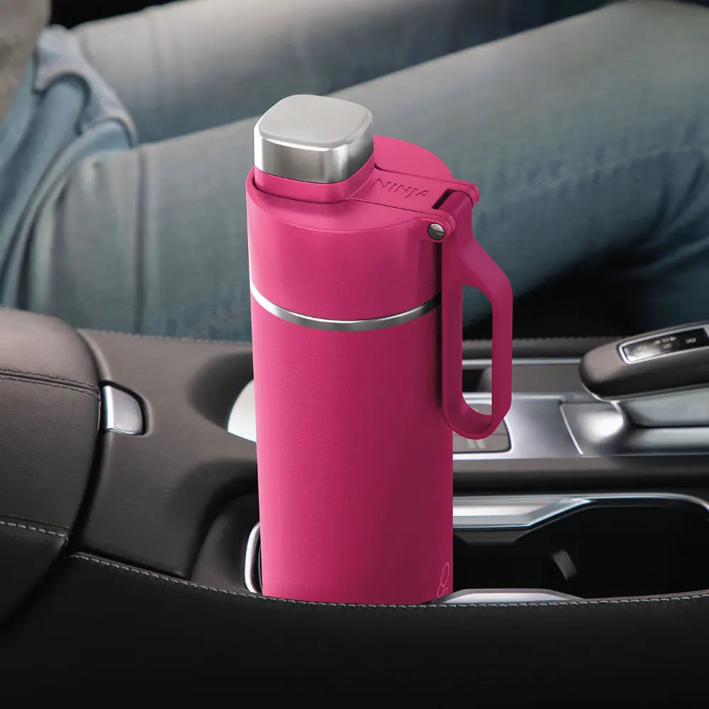 Ninja Thirsti 700ml hot pink insulated travel bottle with stainless steel cap and carrying handle, displayed on car dashboard interior
