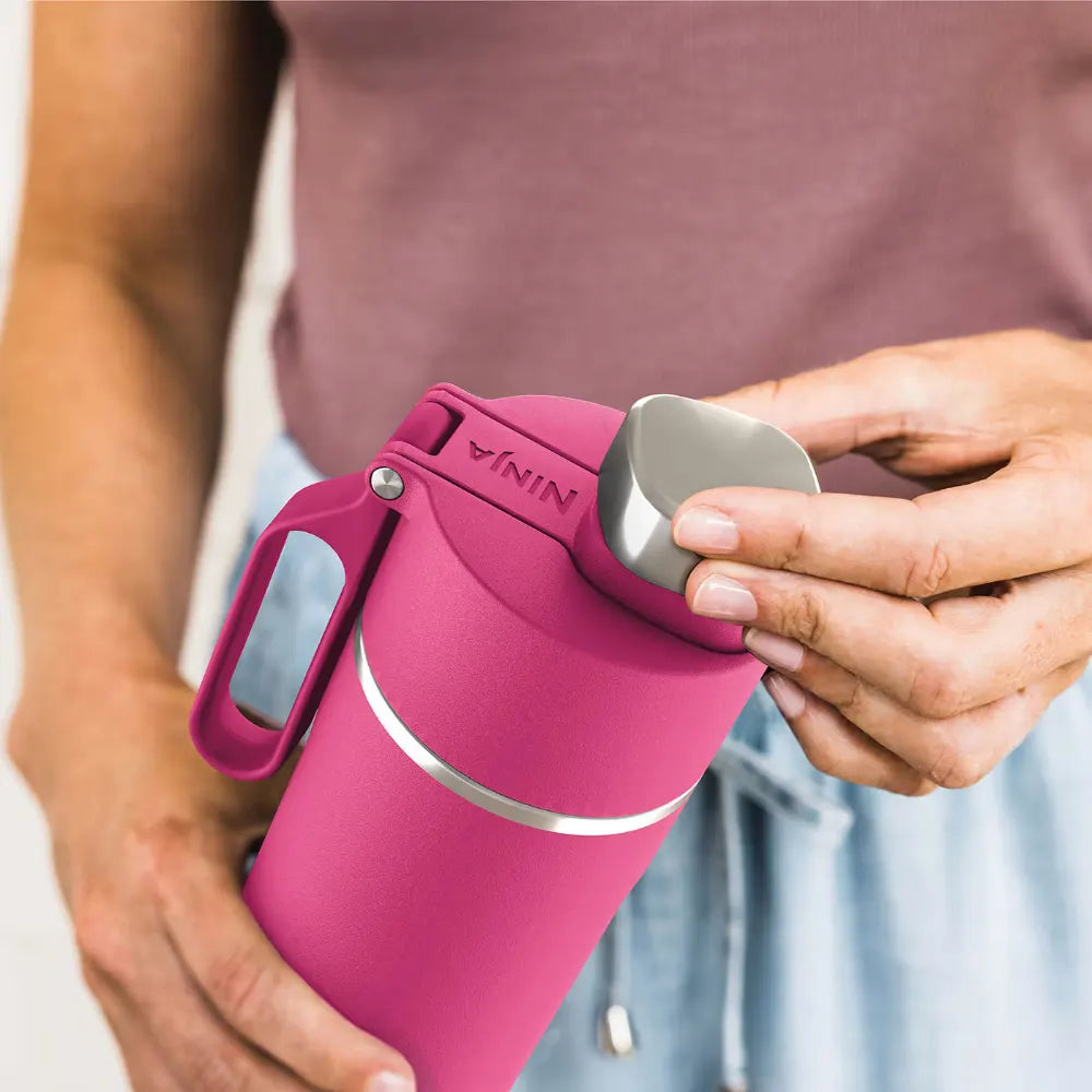 Person holding a hot pink Ninja Thirsti 700ml insulated travel bottle with grey accents and carry handle, displaying the product's design and size in an outdoor setting