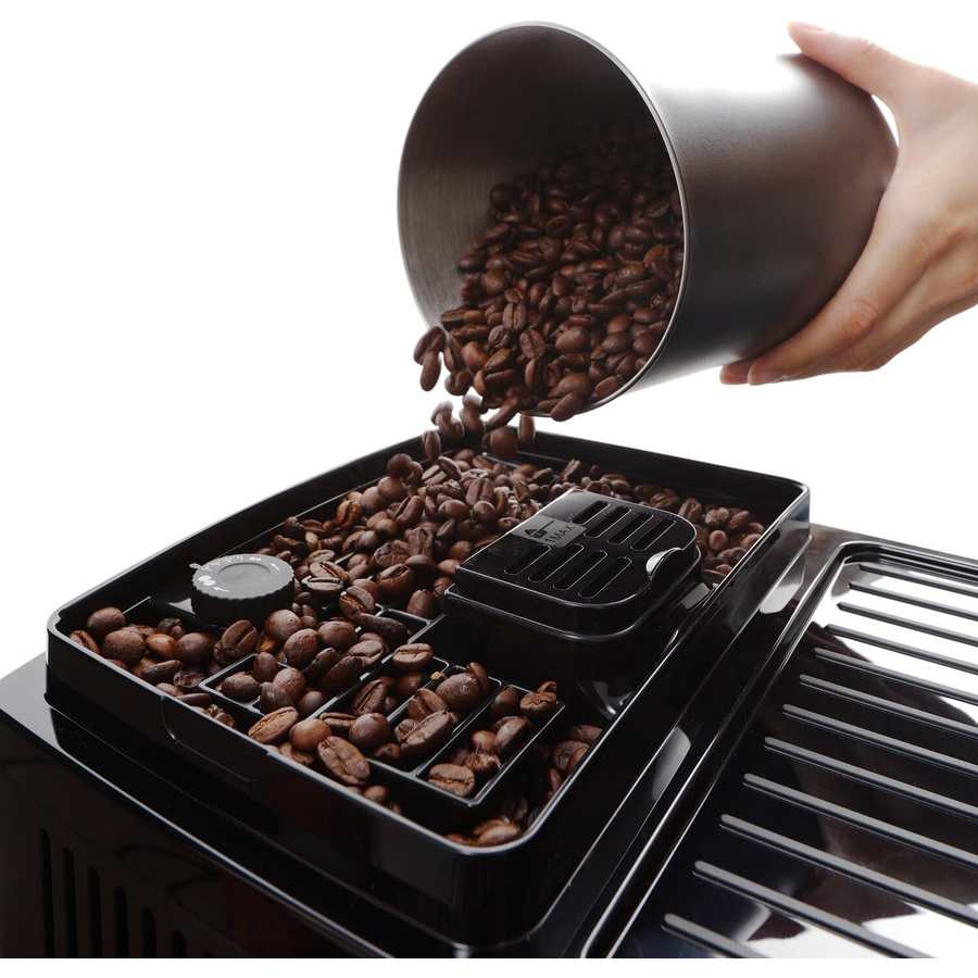 Hand pouring roasted coffee beans from a metal measuring cup into the bean hopper of a black DeLonghi Magnifica Start bean to cup coffee machine with control panel and chrome accents visible