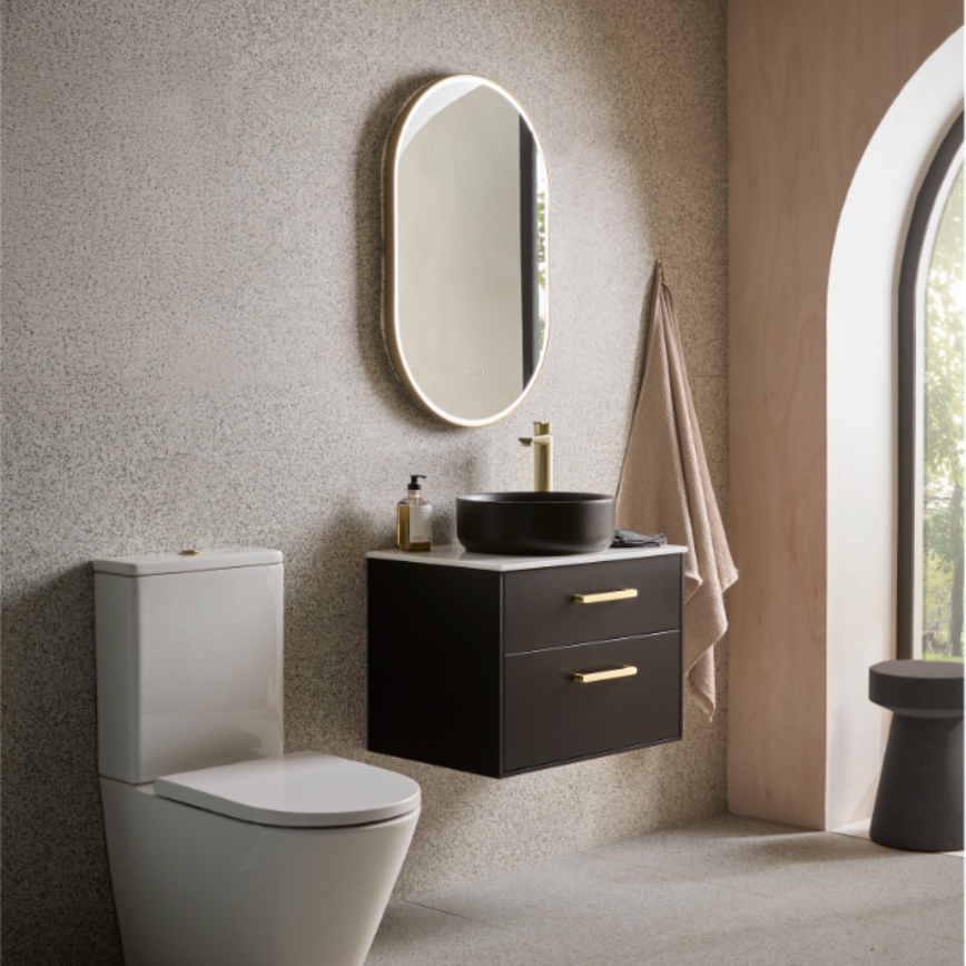 Modern bathroom vanity unit featuring a black wall-mounted cabinet with brass handles, round black vessel sink, illuminated oval mirror with white frame, white toilet, beige textured walls, arched window, and taupe towel in contemporary minimalist design
