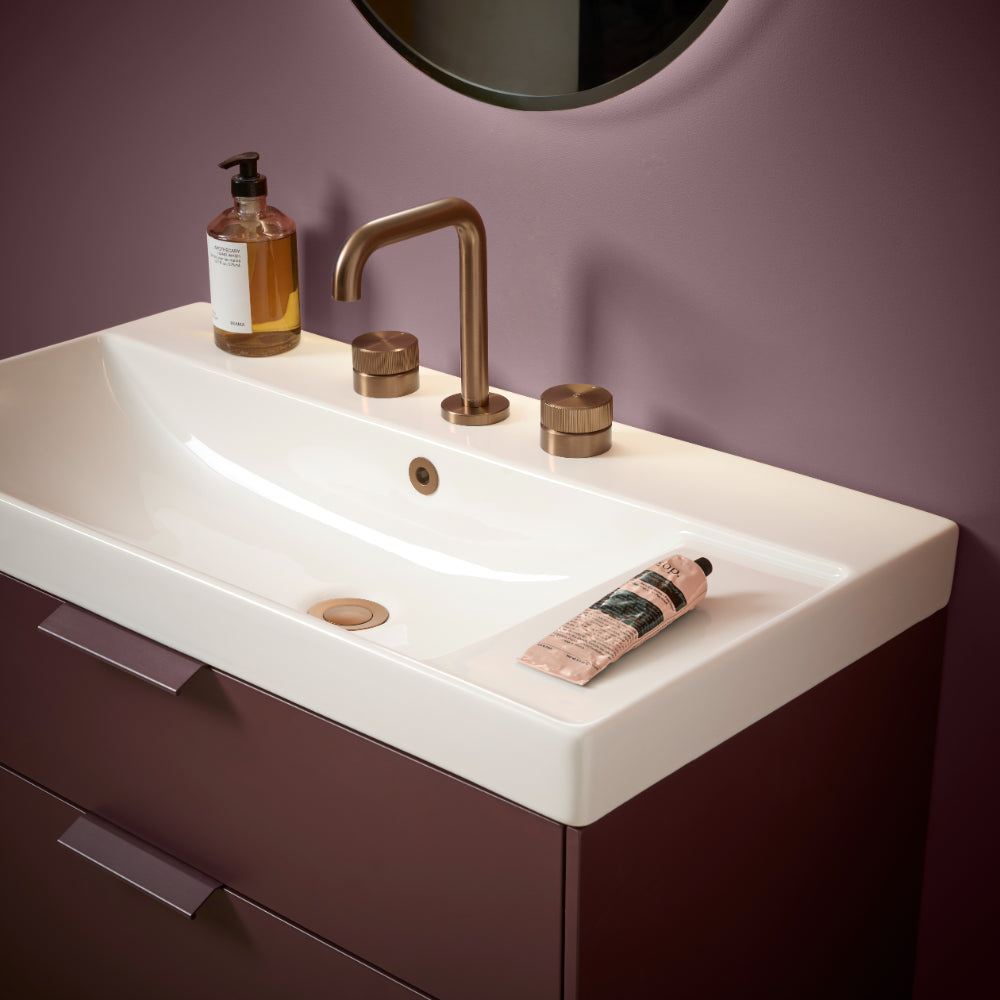 Modern white ceramic rectangular basin sink with brass three-hole tap and knurled handles mounted on burgundy vanity unit, styled with soap dispenser and skincare products against mauve purple wall with circular mirror above