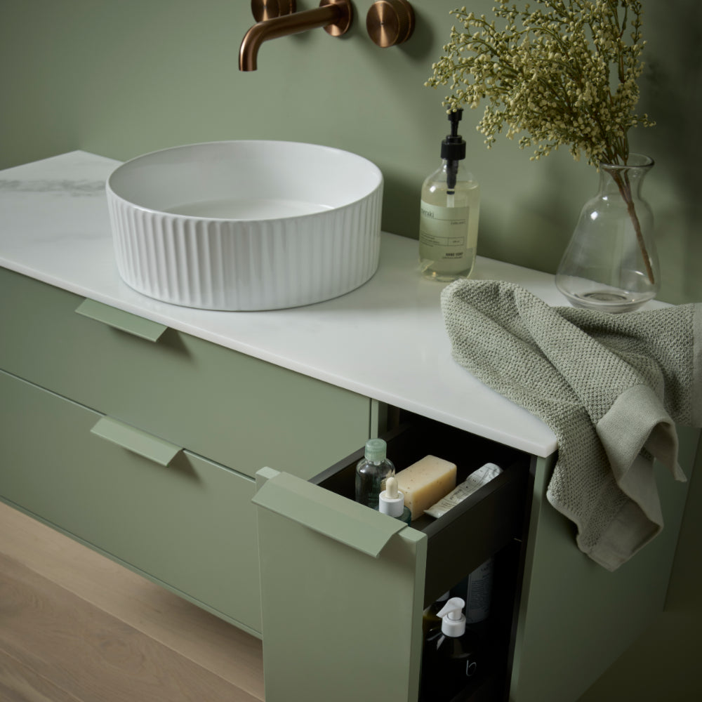 Modern sage green bathroom vanity unit with white ribbed ceramic vessel sink, copper wall-mounted faucet, white countertop, integrated storage drawer, decorative items including glass vase with dried flowers, soap dispenser, folded grey towel, and bathroom accessories