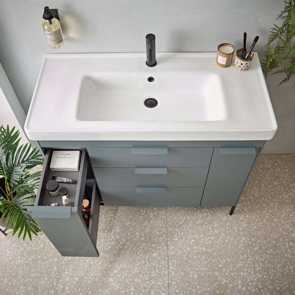 Overhead view of a modern bathroom vanity unit featuring a white rectangular basin with black faucet mounted on a grey-blue cabinet with multiple drawers, styled with bathroom accessories including candles, brushes, and potted plants on the countertop