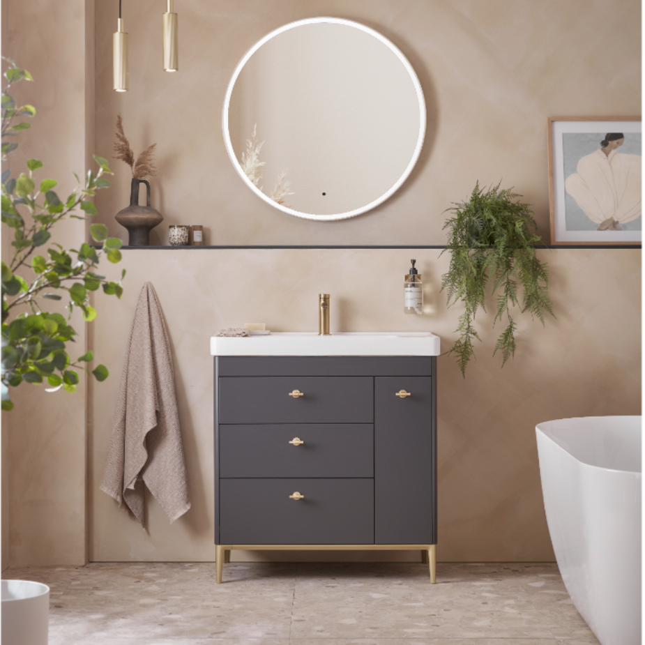 Modern bathroom vanity unit in matt carbon with brass handles and white ceramic basin, styled with circular illuminated mirror, hanging pendant lights, potted plants and neutral beige walls in contemporary spa-like setting