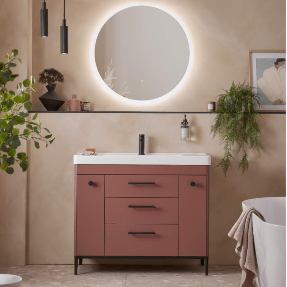 Modern bathroom vanity unit in terracotta with white sink top and black hardware, featuring illuminated circular mirror above, pendant lights, potted plants and accessories in contemporary beige bathroom setting