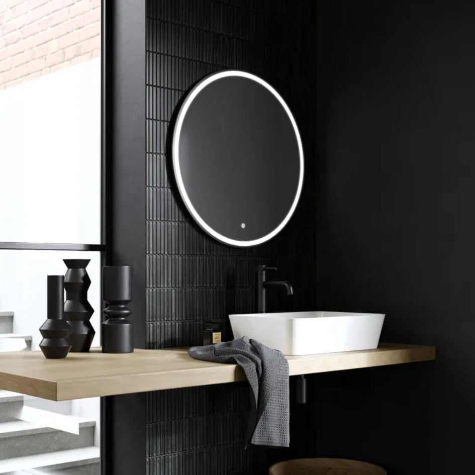 Modern black bathroom with round illuminated LED mirror above white vessel sink on light wood countertop, black wall tiles, and matte black fixtures