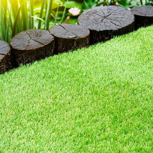 Close-up view of bright green Greenfx Capri artificial grass 28mm pile height with dark wooden log edging and garden plants in background