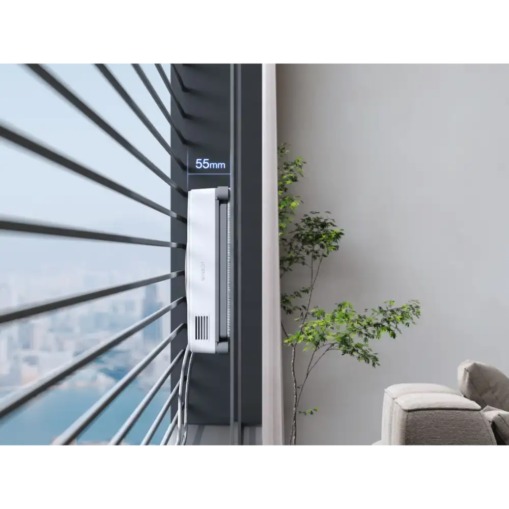 Ecovacs Winbot Mini white robot window cleaner mounted on black window frame with 55mm depth specification, positioned on high-rise apartment window with city skyline view and green potted plant visible indoors