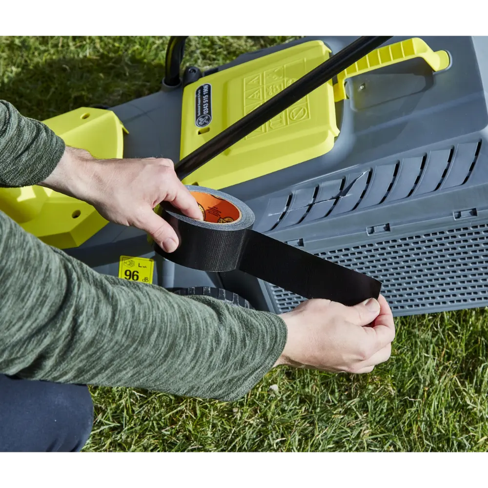 Person applying black Gorilla Tape to yellow and grey tool equipment outdoors on grass, showing the tape's adhesive strength and durability in practical use