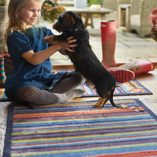 Young girl sitting on a colorful striped Hug Rug runner mat playing with a black dachshund dog in a sunny outdoor patio setting