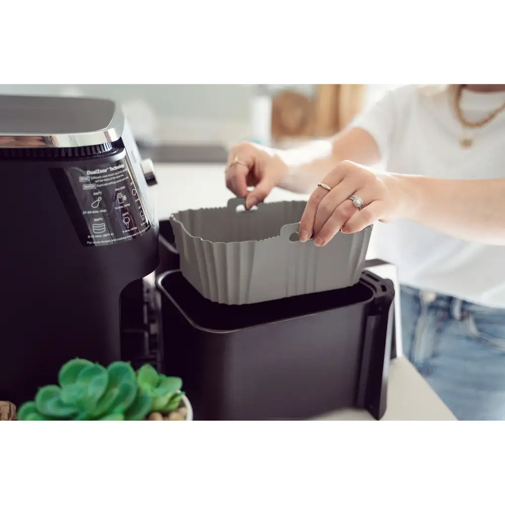 Woman in white shirt placing gray silicone air fryer liner into black air fryer basket next to air fryer unit with succulent plant visible in foreground