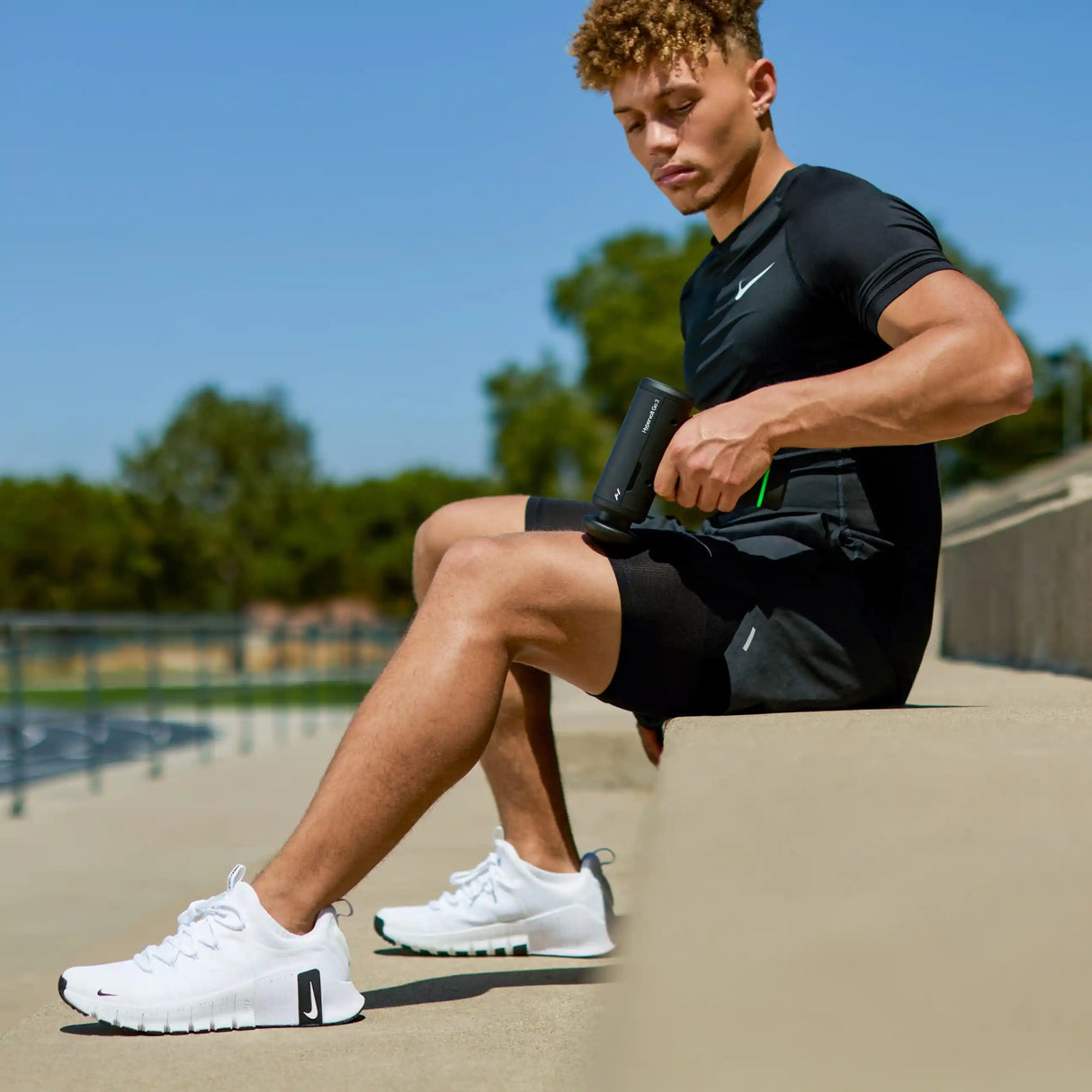 Young male athlete in black Nike sports shirt and shorts wearing white sneakers sits poolside using a black Hyperice Hypervolt Go 3 massage gun on his arm for muscle recovery