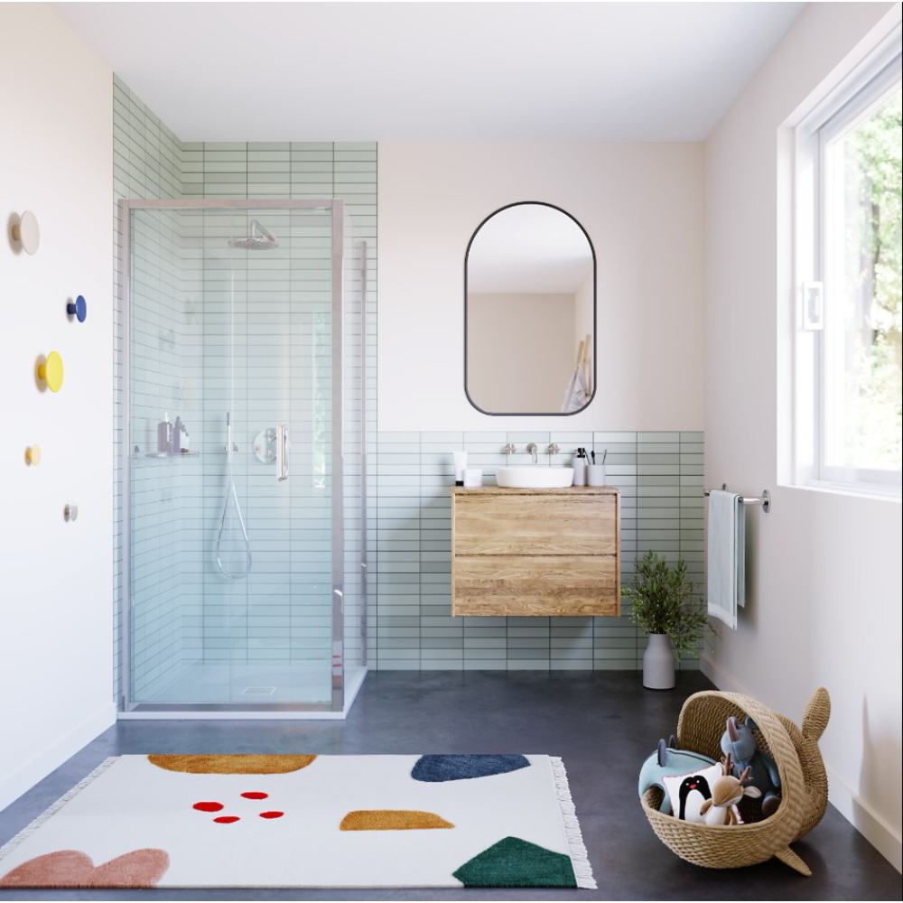 Modern bathroom featuring a Flair ILI chrome bifold shower door with light blue subway tiles, wooden vanity with white vessel sink, black-framed oval mirror, dark grey floor, colorful geometric bath mat, and natural lighting from window