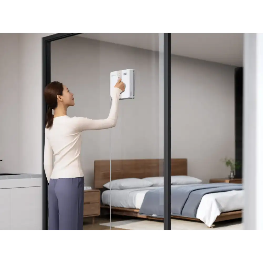 Woman in cream long-sleeve top and navy pants operating Ecovacs Winbot Mini white robot window vacuum on bedroom mirror, with wooden bed frame and gray bedding visible in reflection