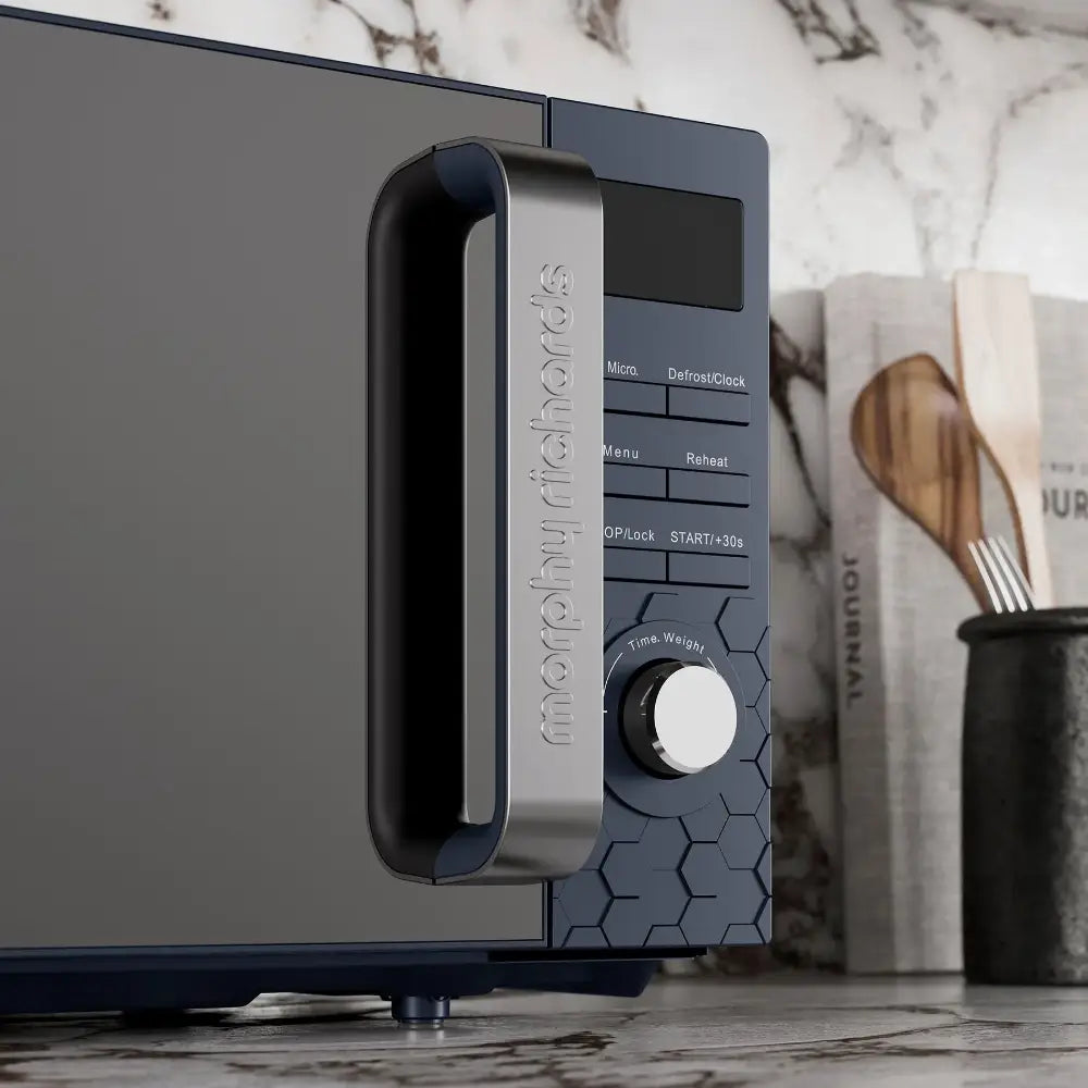 Morphy Richards Cassini Digital Microwave 20 Litre 700W in Outer Space blue with stainless steel handle, featuring control panel with buttons for Defrost/Clock, Menu, Reheat, Stop/Lock and START/+30s, plus Time Weight dial, displayed on marble countertop with kitchen utensils in background
