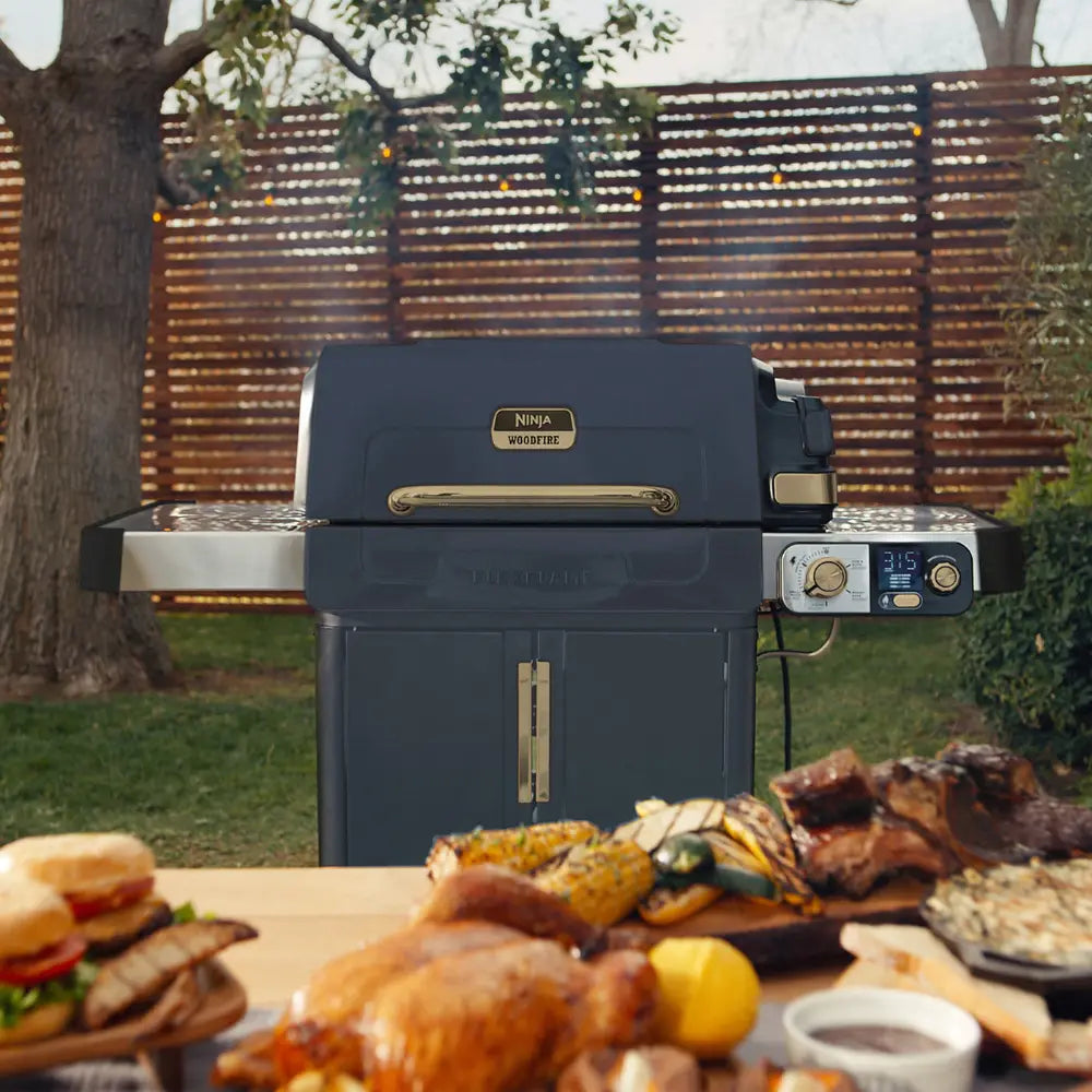 Ninja FlexFlame gas BBQ grill in navy blue with brass accents set in a garden with wooden slatted fence, displaying grilled food including corn, meat and burgers on a wooden table in foreground