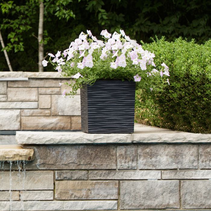 Slate grey rippled garden planter with white and pale pink trailing petunias and green foliage, positioned on a natural stone wall with water feature and hedges in background