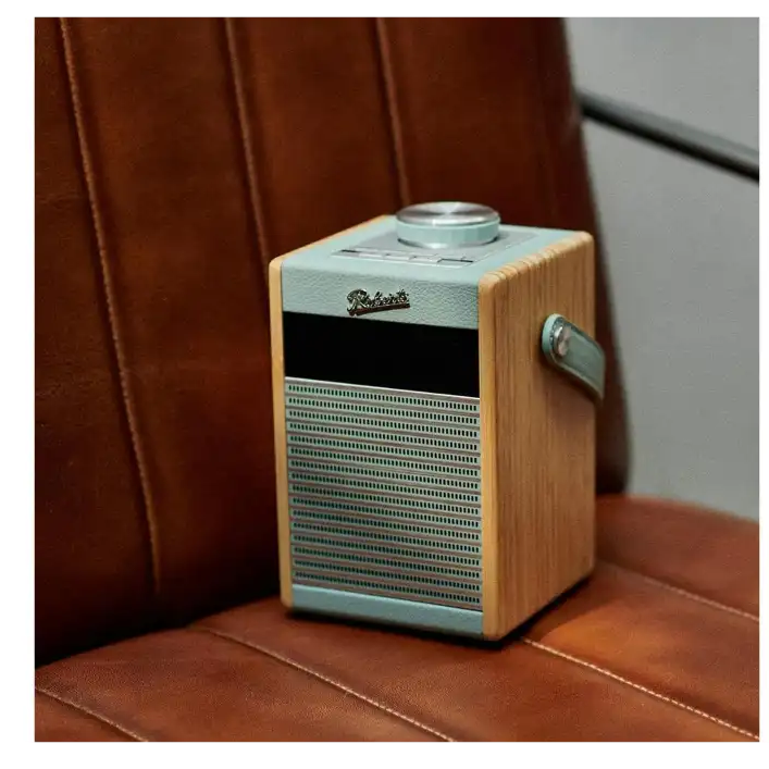 Roberts Rambler Midi DAB+ FM radio with Bluetooth in duck egg blue and natural wood finish, sitting on a brown leather surface with a burgundy leather chair in the background