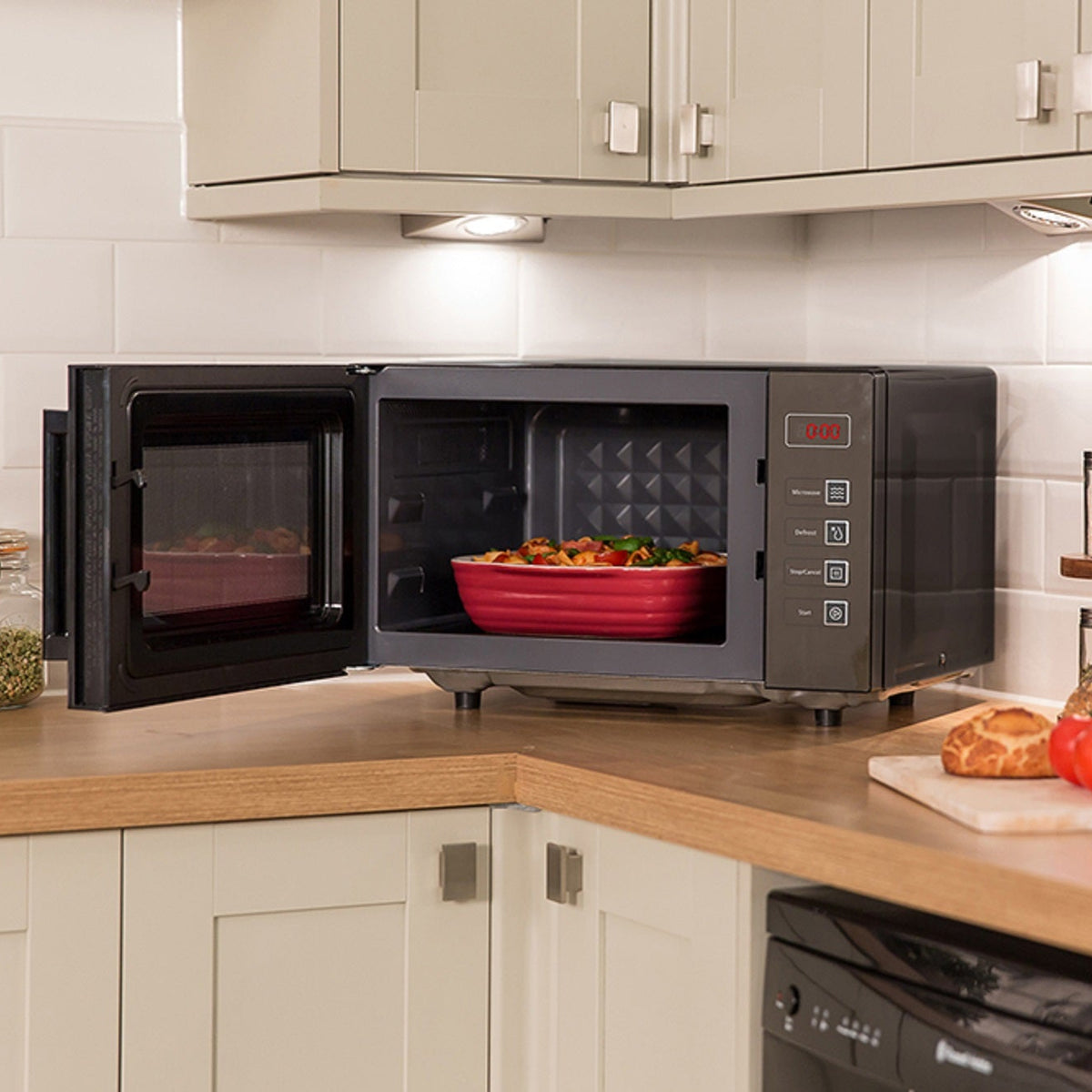 Russell Hobbs black 23 litre flatbed microwave with red dish of food inside, displayed on wooden kitchen countertop with cream cabinets, white tile backsplash, fresh tomatoes and bread on cutting board, and glass storage jars with herbs and spices