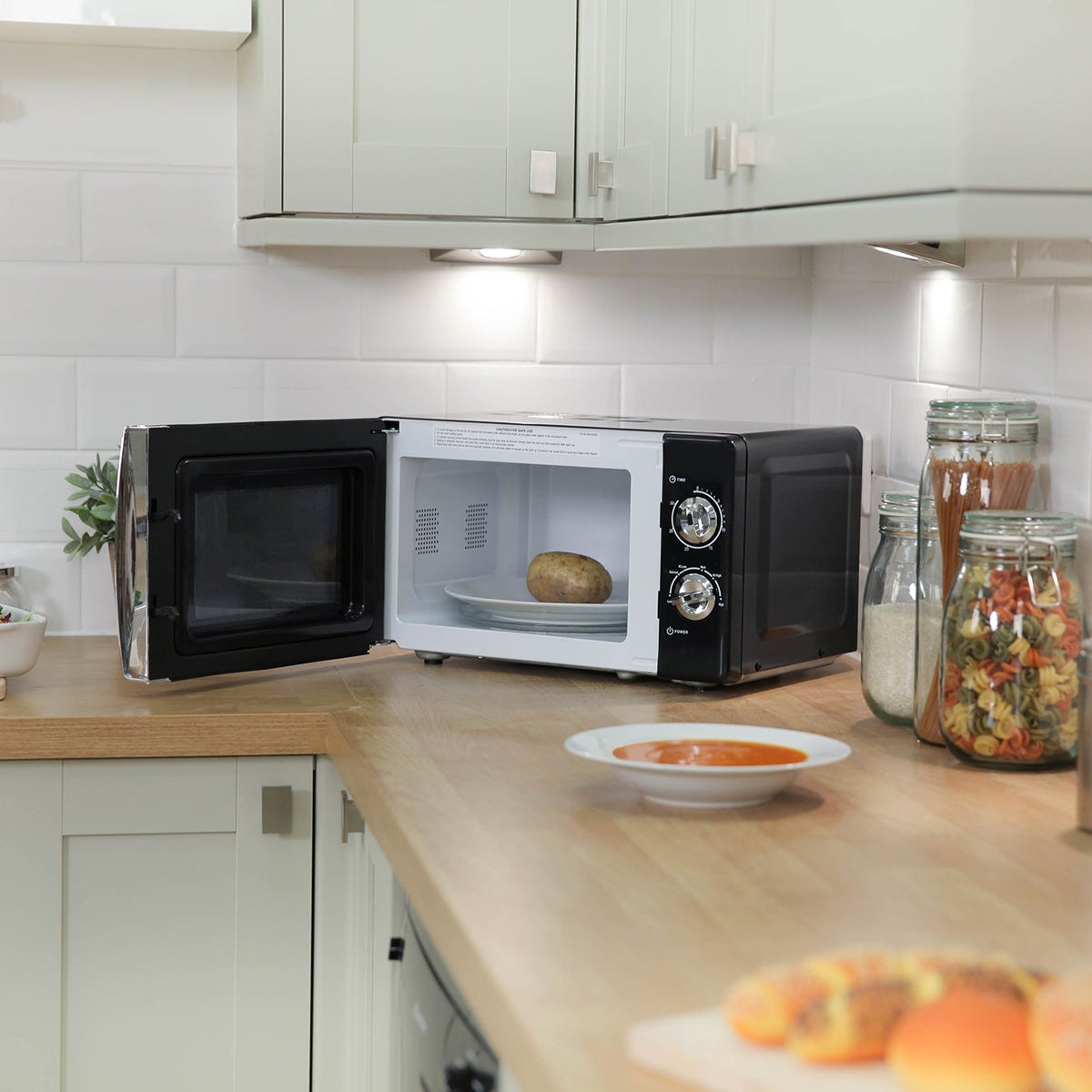 Russell Hobbs 700w black microwave with open door showing potato inside, positioned on wooden kitchen countertop beside glass storage jars, white subway tile backsplash, and light green cabinets above