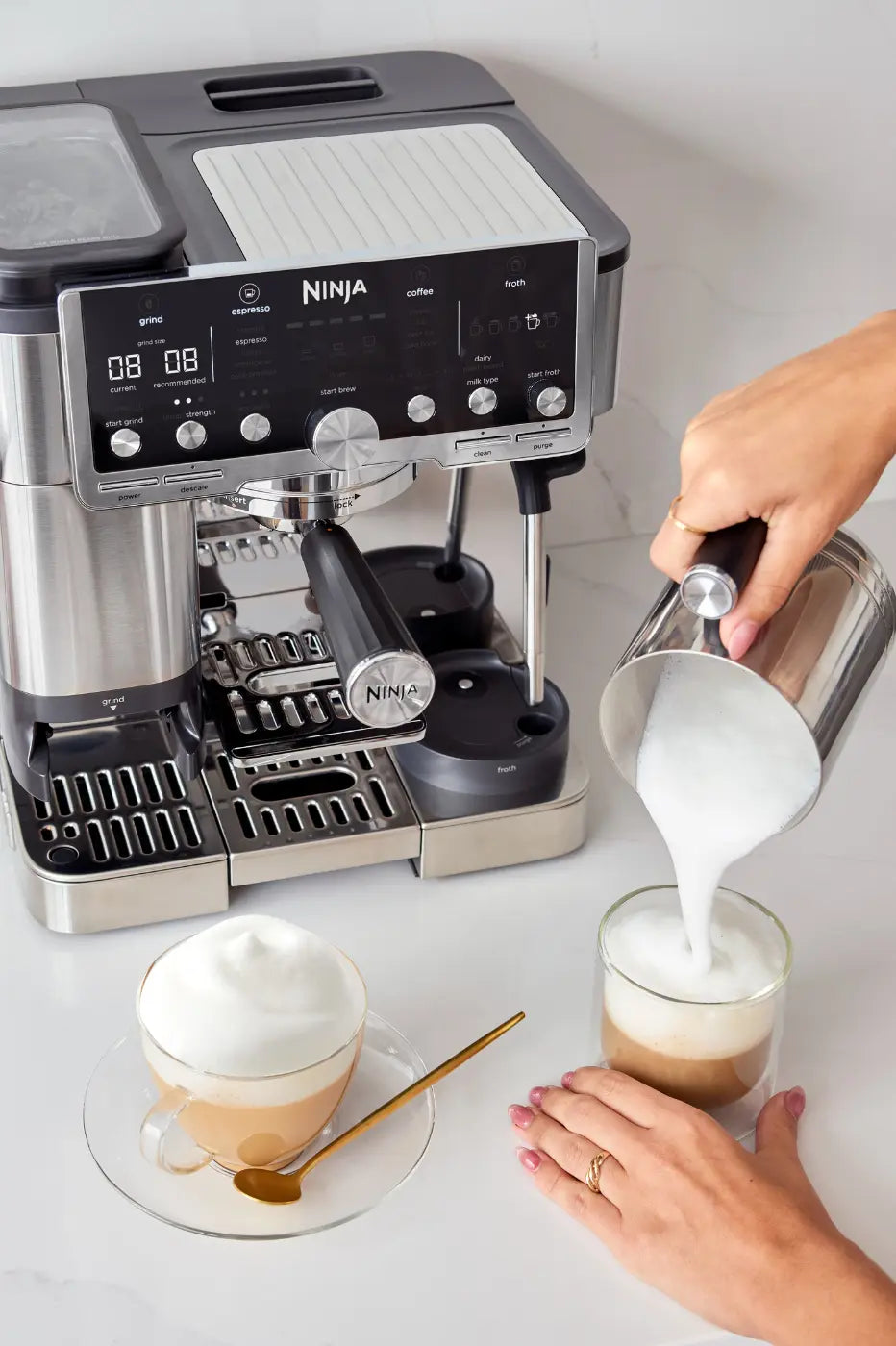 A stainless steel and black Ninja espresso machine on a light marble countertop, with a person pouring milk from a metal pitcher into a glass of latte. Another cup of topped latte with foam sits on a saucer with a gold spoon nearby.
