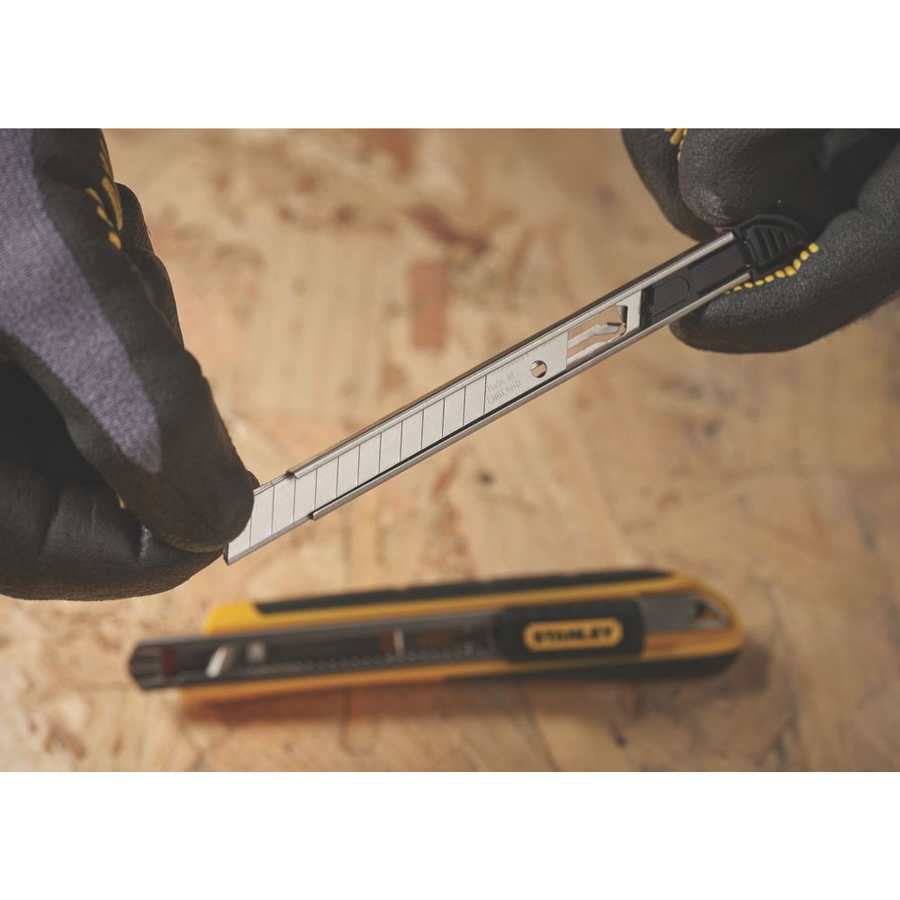 Person in black work gloves holding a Stanley snap-off utility knife blade with metal casing above a Stanley utility knife on a concrete surface