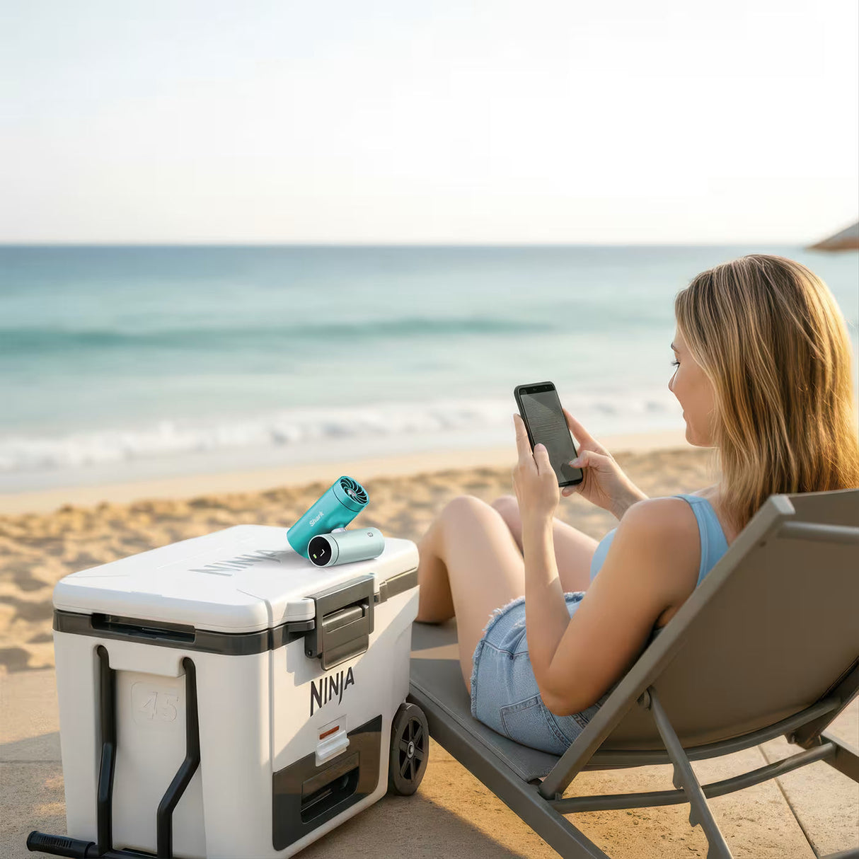 Woman sitting on a beach chair using a smartphone next to a white cooler with 'Ninja' branding.