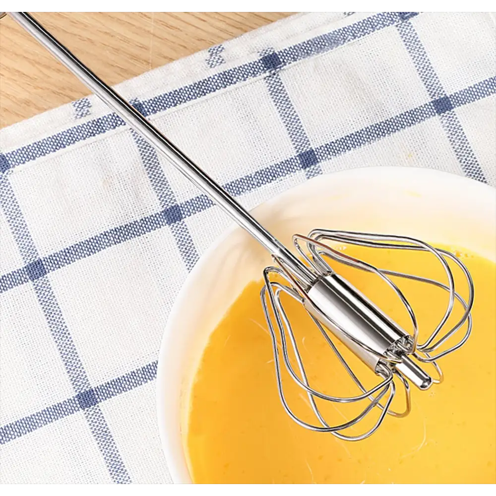 Manual push spin whisk with stainless steel handle resting in bowl of yellow liquid on blue and white checkered kitchen towel next to wooden floor