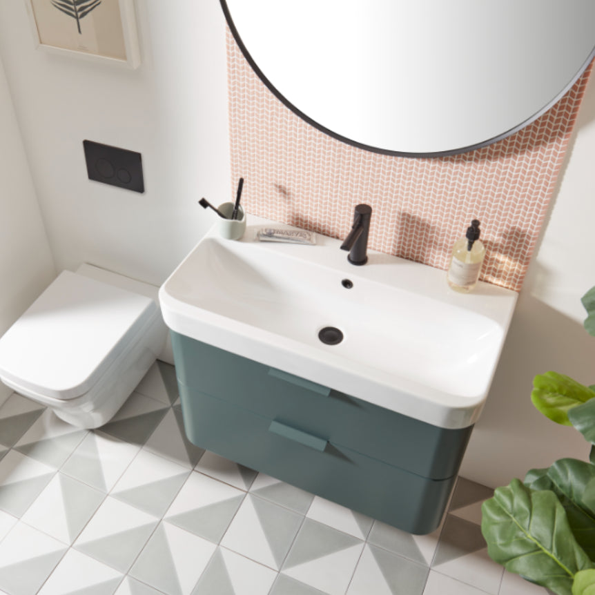 Modern bathroom vanity unit with Derwent Blue cabinet base and white rectangular basin, black tap and accessories, paired with round black-framed mirror above peachy-pink geometric tile backsplash, white geometric floor tiles, matching white toilet, and green potted plant