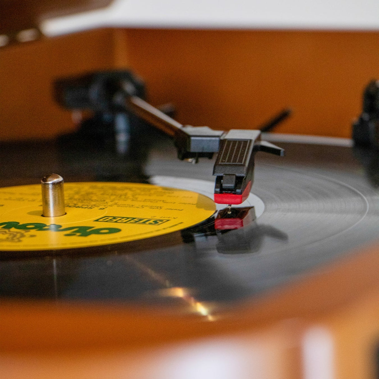 Close-up of a Lenco Classic Phono turntable with yellow vinyl record and red tonearm cartridge, showing the needle on the spinning platter against a warm brown wooden surface with blurred turntables in the background