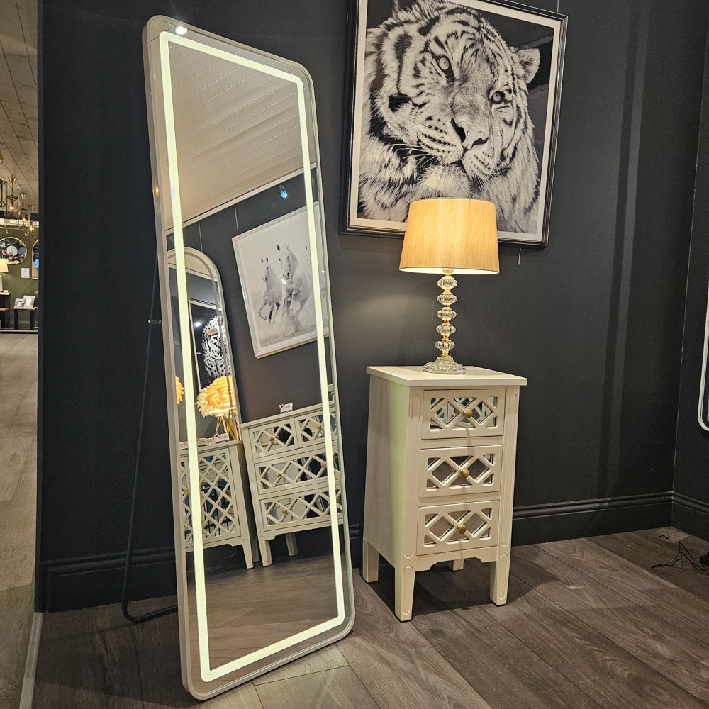 Tara Lane Modena LED Cheval Mirror in white with illuminated frame displayed in showroom next to cream nightstand with geometric patterns and brass table lamp, against black wall with tiger artwork