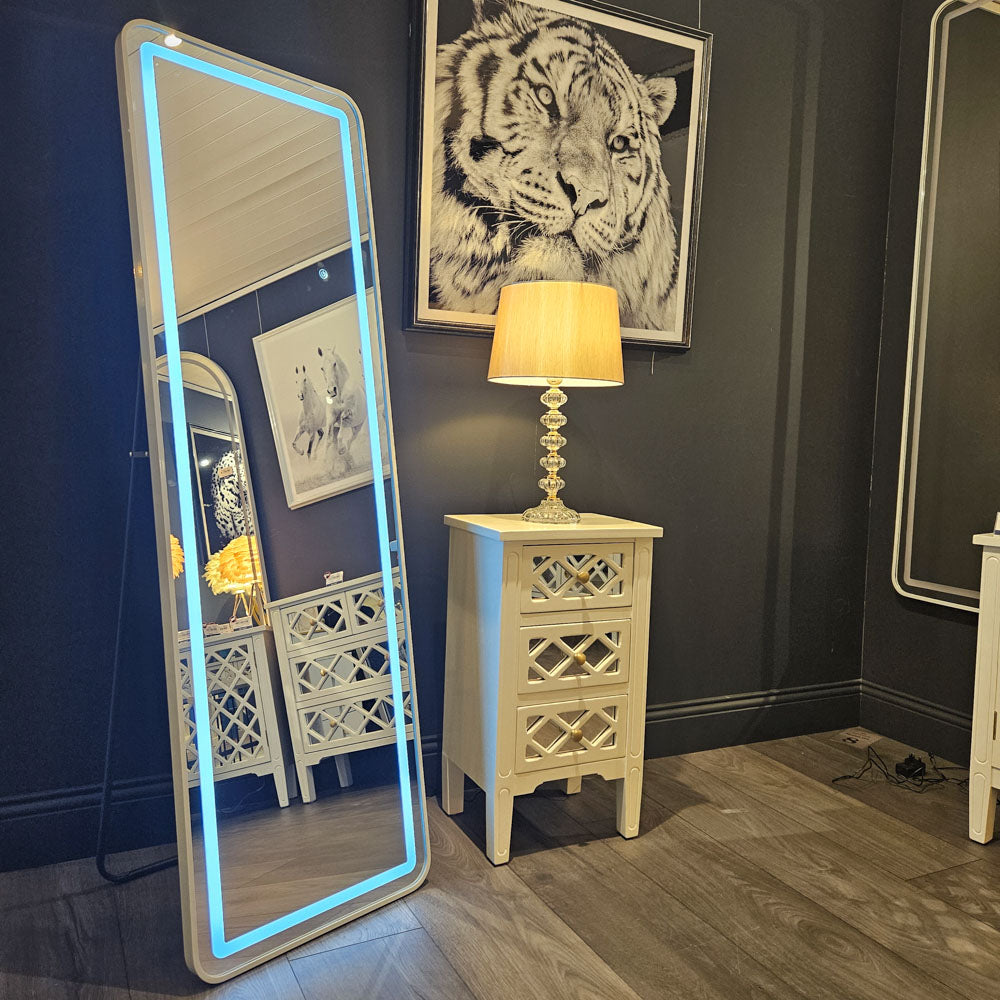Tara Lane Modena LED Cheval Mirror with white frame and blue neon border in modern showroom setting, displayed next to cream wooden nightstand with lamp and tiger artwork on dark wall