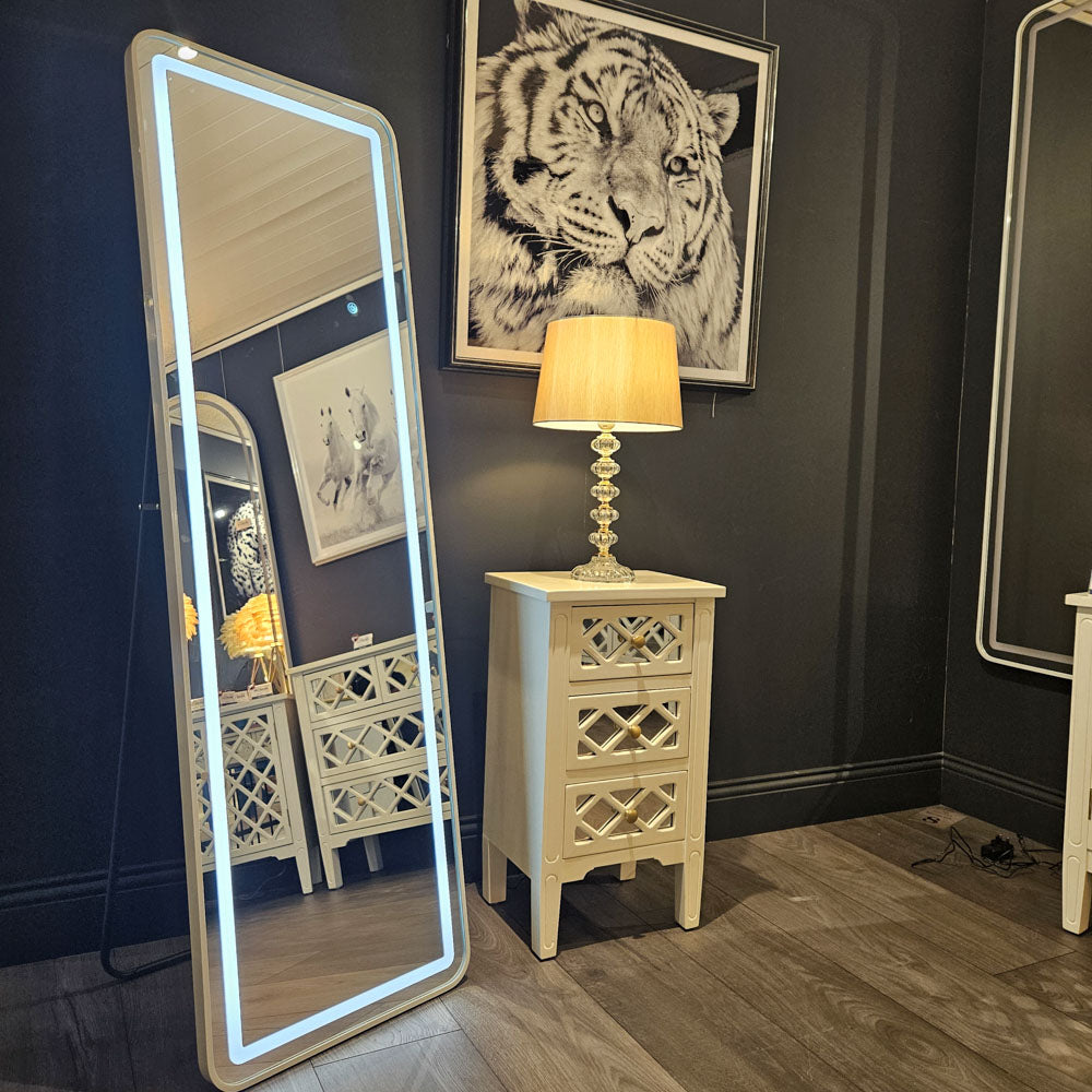 Tara Lane Modena LED cheval mirror with white frame and blue neon lighting displayed in a showroom setting next to a cream wooden nightstand with lamp and tiger artwork on dark wall