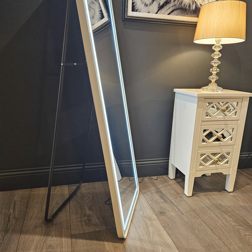 Tara Lane Modena LED Cheval Mirror white frame leaning against dark wall beside cream nightstand with lamp in modern bedroom setting