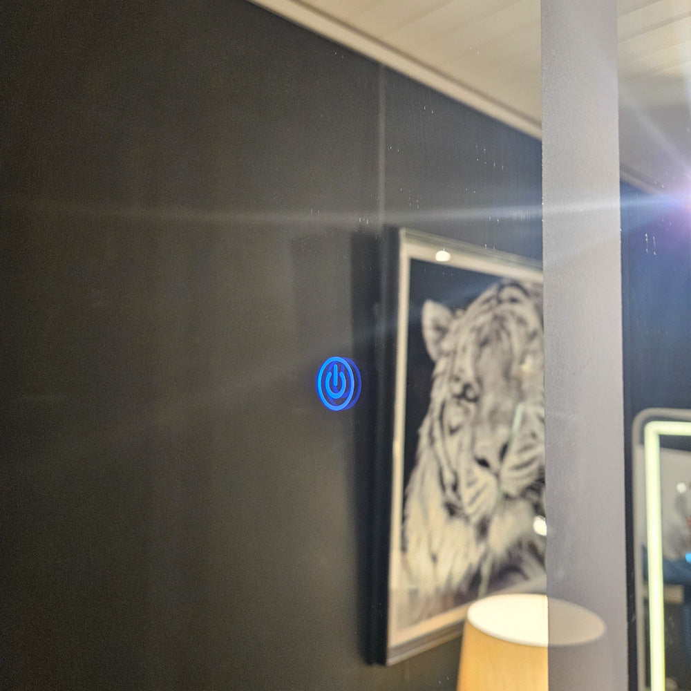 Blue power button illuminated on dark glass surface of modern LED mirror with white frame and framed artwork visible in reflection