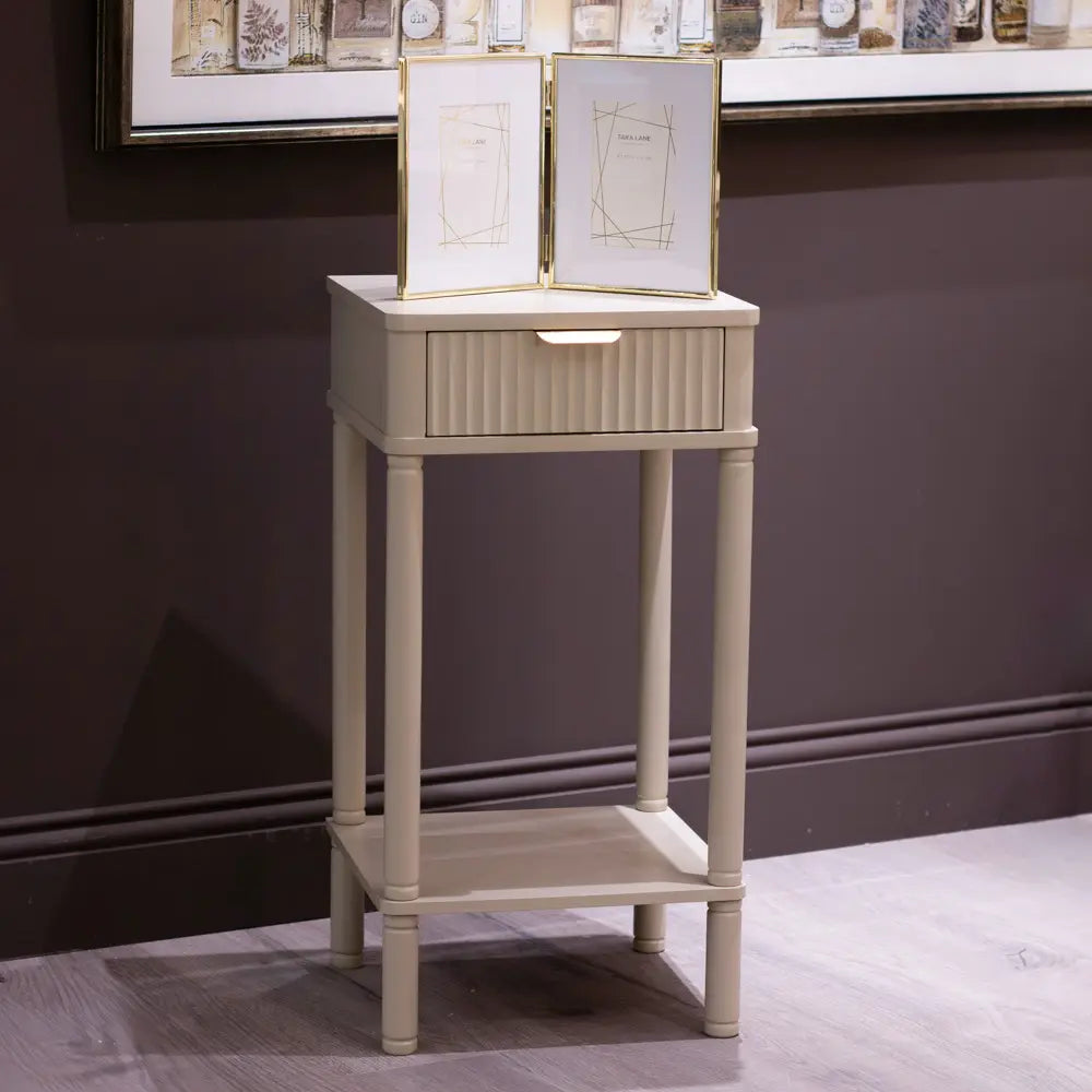 Tara Lane Parson cream oyster-colored accent table with single drawer and open lower shelf, featuring fluted drawer front and cylindrical legs, displayed in showroom with framed artwork on wall behind
