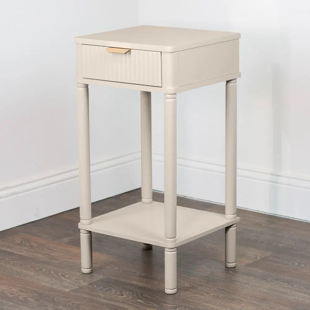 Cream colored wooden accent table with single drawer, cylindrical legs and lower shelf on dark wood floor against white wall