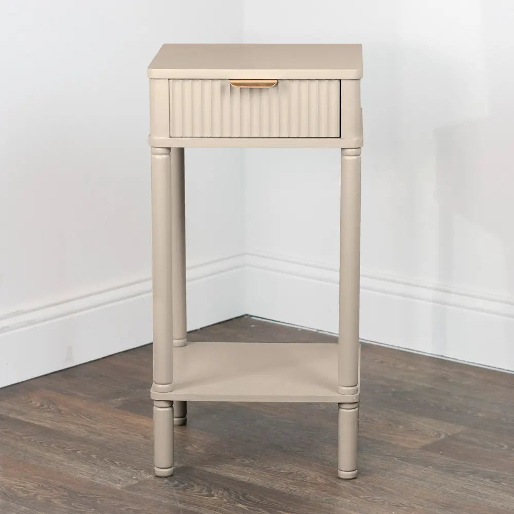 Cream colored wooden accent table with one drawer and open shelf, featuring vertical reeded detailing on drawer front and turned legs, displayed against white baseboards on dark hardwood flooring