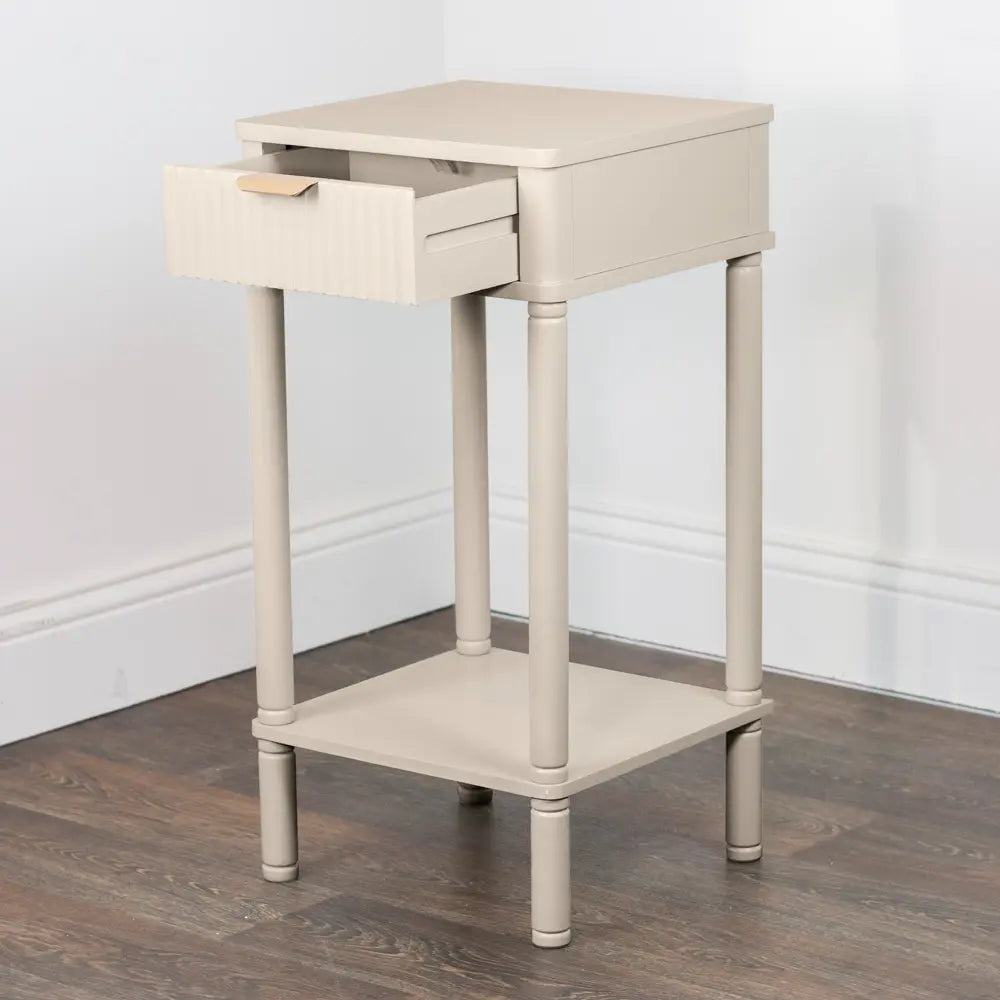 Cream-colored wooden accent table with one open drawer, lower shelf, and turned legs on dark hardwood floor against white wall