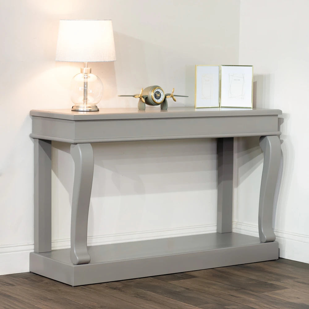 Mink grey Tara Lane Scroll Console Table 130cm with curved legs, featuring a table lamp, decorative airplane clock, and framed artwork on top, displayed in a bright interior setting with wooden flooring