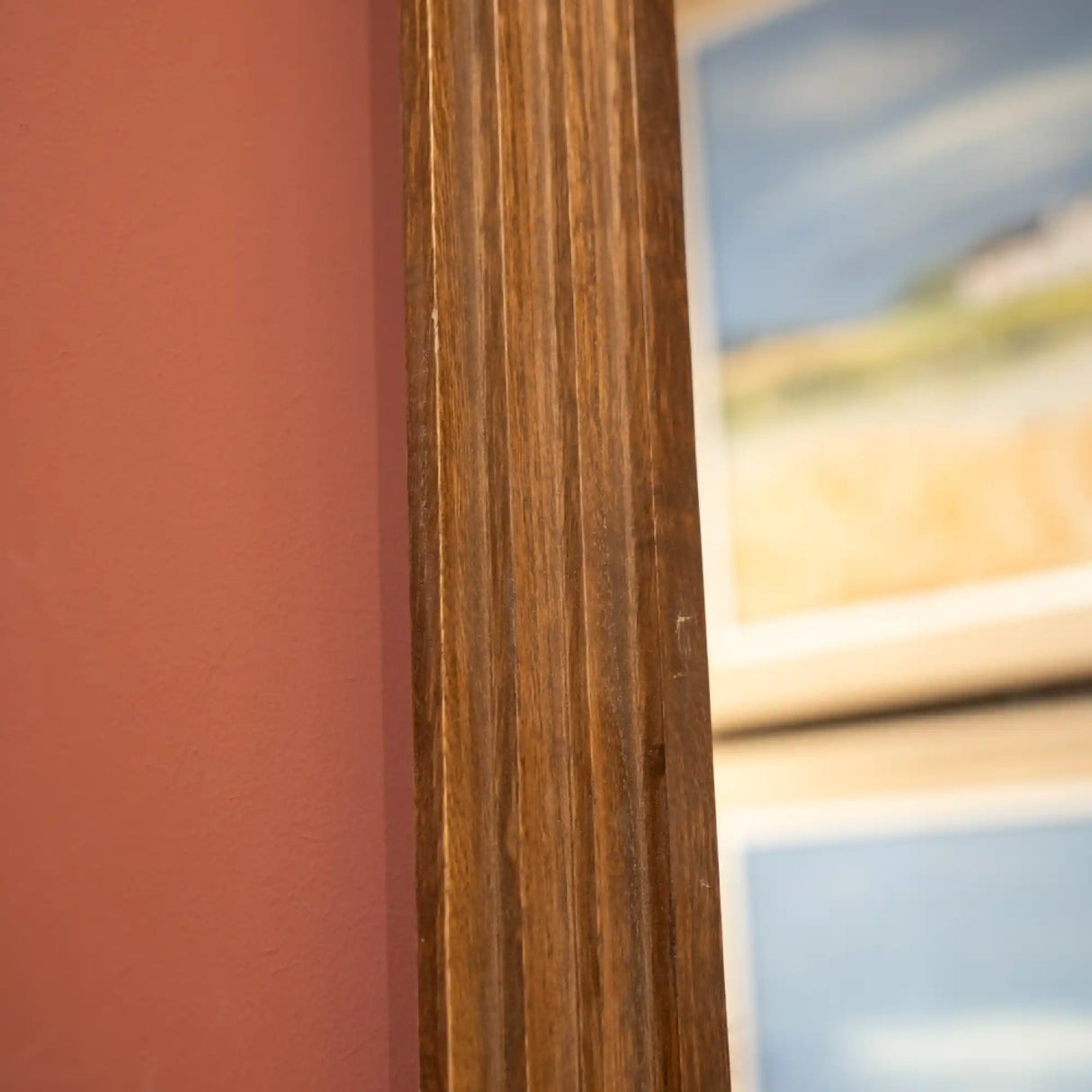 Tall pill-shaped dressing mirror with natural wood frame featuring vertical grain detail, positioned against coral pink wall with blurred landscape artwork visible in reflection