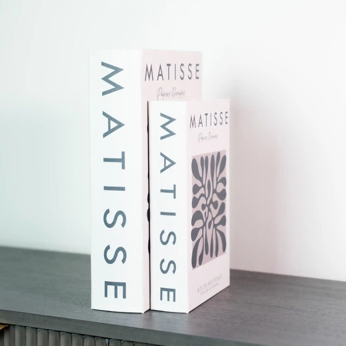 Two white paperback books with gray lettering displaying "Matisse" by Tara Lane standing upright on a dark surface, featuring minimalist design with pink accents and decorative botanical motifs on the spines