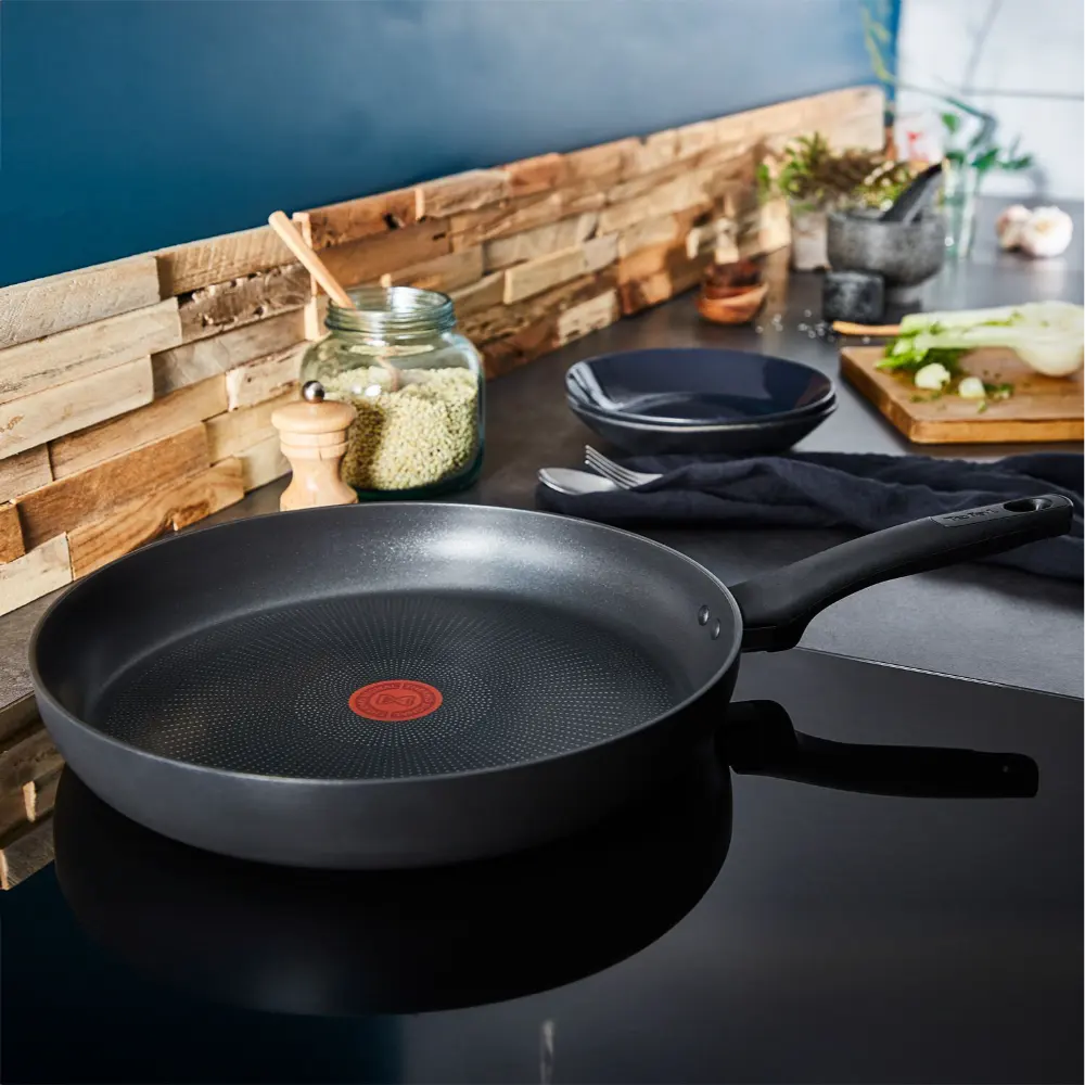Tefal Ultimate 30cm black non-stick frying pan displayed on dark countertop with wooden backsplash, featuring red circular logo, alongside black bowls, wooden cutting board with vegetables, glass jar, and kitchen accessories in modern kitchen setting