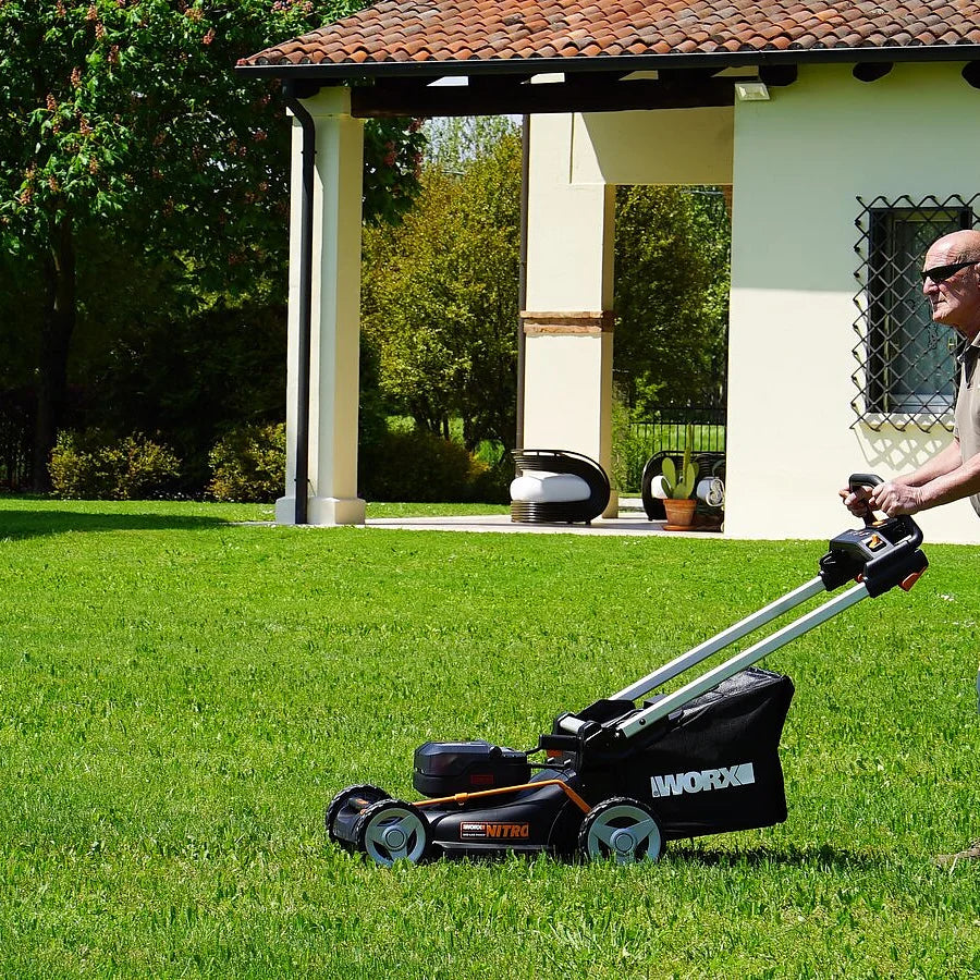 Man using Worx 40V Brushless Self Propelled Battery Lawn Mower with 46cm cutting deck on green lawn in front of modern white house with terracotta roof and black pergola