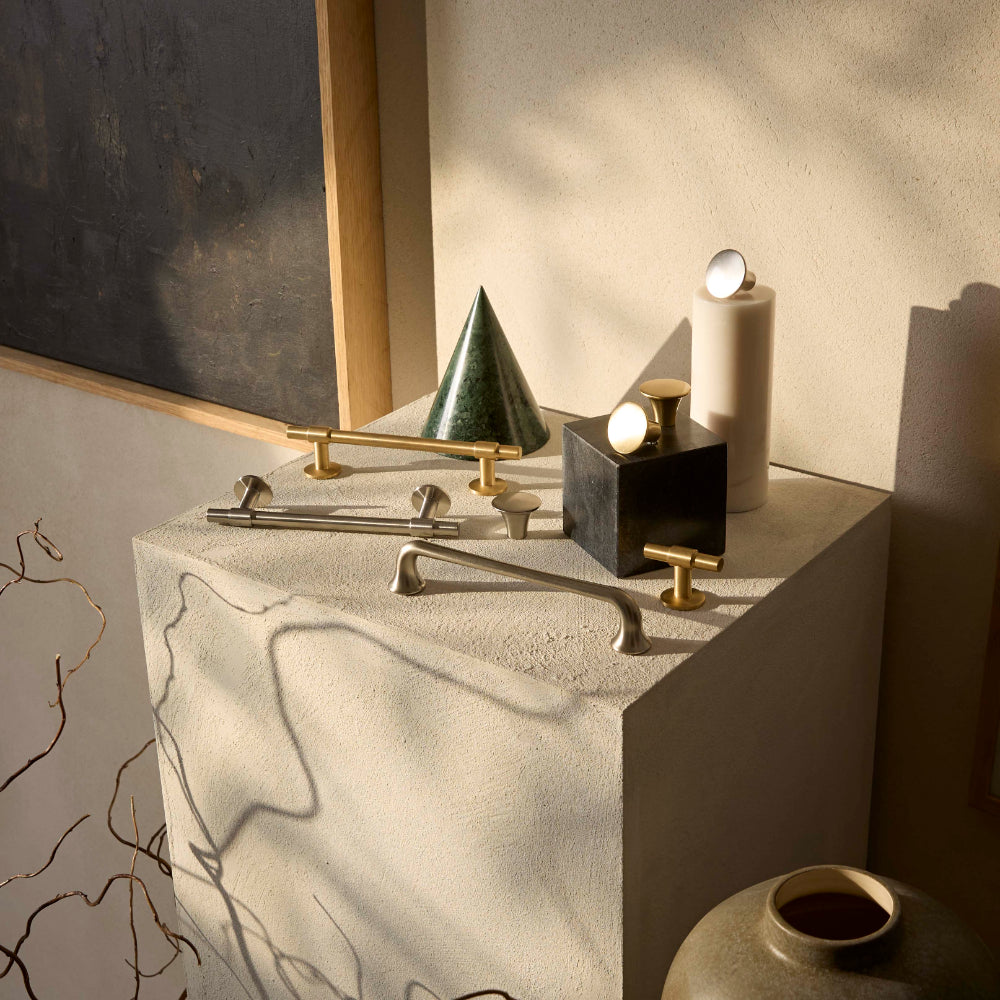 Styled bathroom vanity display featuring beige stone sculptural forms with brass hardware and minimalist fixtures, including cone-shaped green marble object, cylindrical white ceramic vessels, and small brass pull handles arranged on sandy textured surface with warm natural lighting